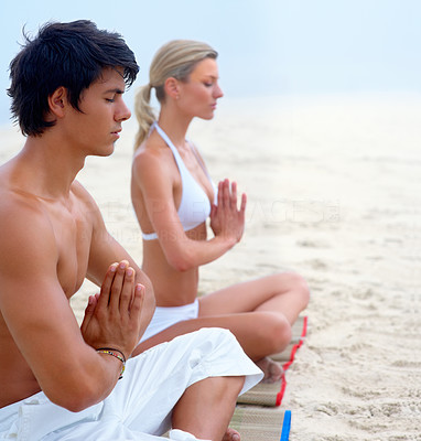 Young man and woman sitting at the beach practicing yoga together with their hands joined