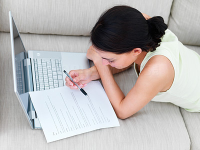 Young woman lying on sofa using laptop, reading paper