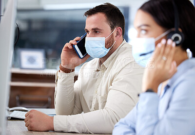 Buy stock photo Shot of a young businessman sitting in the office with his colleague and wearing a face mask while using his cellphone