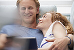 A smiling young couple resting on their couch while using a digital tablet
