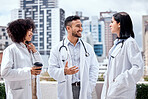 Two female doctors and a male colleague standing on the balcony outside a hospital smiling and having a conversation. mixed man and woman doctors talking to a caucasian female doctor while having a cup of coffee