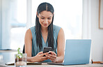 Happy young mixed race businesswoman smiling while using a phone and working on a laptop sitting in a chair in an office. One hispanic female boss using social media on a phone while sending an email