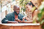 Interracial couple using a phone together while sitting outside at a cafe. Mixed race woman showing an african american man a message on her phone sitting at a restaurant