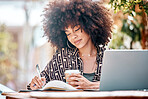 Young happy stylish mixed race businesswoman with a curly afro writing in a diary drinking coffee and working on a laptop sitting outside at a cafe. Hispanic female student studying at a restaurant