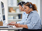 Focused young mixed race female call centre telemarketing agent writing notes and talking on a headset while working on a computer in an office. Serious african american businesswoman consultant operating helpdesk for customer service support