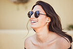 Close up of cheerful mixed race woman wearing sunglasses and laughing while standing outside on sidewalk. Joyful young brunette woman looking stylish in a off-shoulder top on a summer day