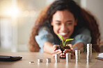 Young happy hispanic woman holding a plant growing out of soil in her hands with money coins on different levels on a table. Symbolises money growing, investment and banking