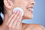 A beautiful smiling mixed race woman using a cotton pad to remove makeup during a selfcare grooming routine. Hispanic woman applying cleanser to her face against blue copyspace background