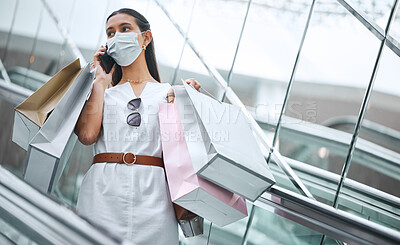 Buy stock photo One young mixed race woman wearing a face mask and talking on a cellphone while on an escalator after a shopping spree. Fashionable hispanic carrying retail bags after buying in a mall during Covid