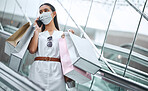 One young mixed race woman wearing a medical face mask and talking on a cellphone while on an escalator after a shopping spree. Fashionable hispanic carrying retail bags after buying in a mall during Covid-19 pandemic