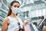 Portrait of a young mixed race woman wearing a face mask and holding a cellphone while on an escalator after a shopping spree. Fashionable hispanic carrying retail bags after buying in a mall during Covid