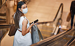 Portrait of a young mixed race woman wearing a face mask and holding a cellphone while on an escalator after a shopping spree. Fashionable hispanic carrying retail bags after buying in a mall during Covid