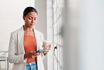 Young mixed race businesswoman using her phone while drinking a coffee alone in an office at work. Confident hispanic businessperson using social media on her cellphone while holding a coffee cup on a break while standing at work