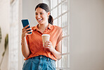 Young happy mixed race businesswoman using her phone while drinking a coffee alone in an office at work. One cheerful hispanic businessperson using social media on her cellphone while holding a coffee cup on a break while standing at work