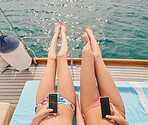 Two friends sunbathing on boat during holiday cruise together using cellphones to send text messages. Two women lying on boat using smartphones and sunbathing in bikinis