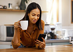 Young mixed race woman drinking a cup of coffee while using her phone alone in the morning in the kitchen. Content hispanic female smiling and enjoying a cup of tea while using social media on her phone at home