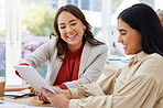 Team of two smiling business women using a digital tablet for a brainstorm meeting in the office. Happy confident ethnic professionals using technology while talking and planning a marketing strategy
