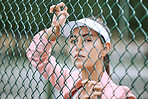 Close up of a female athlete leaning against a wire fence. Young hispanic tennis player posing on a tennis court wearing a white visor and pink jacket